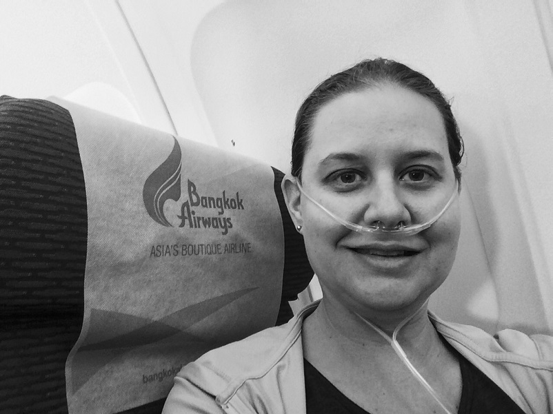 Woman smiling wearing a oxygen hose sitting on a plane in front of a seat cushion that says "Bangkok Airways"