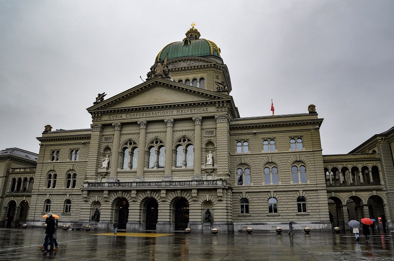 People with umbrellas walking in front of Bern Parliament building on a grey day in Switzerland