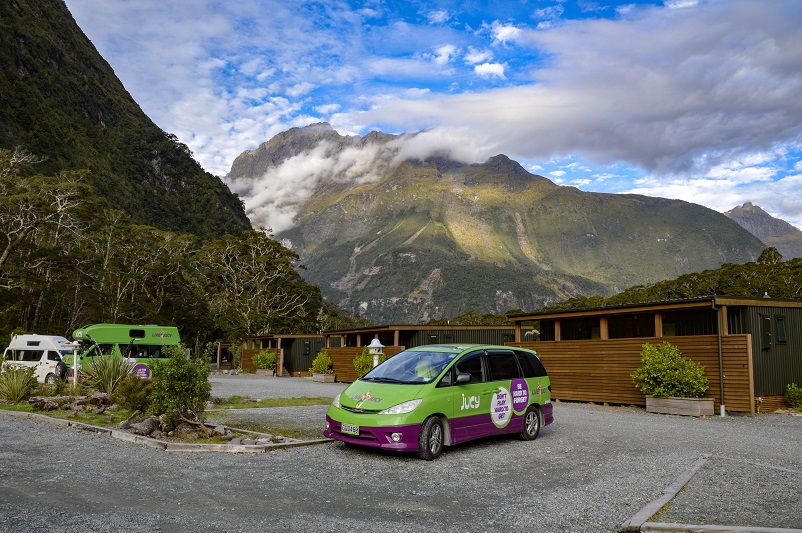 Vans parked in parking lot in front of Milford Sound lodge with backdrop of mountains, NZ