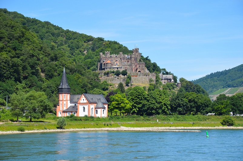 Pink, white and black building in front of a castle on a hill as seen from a Rhine River cruise in Germany