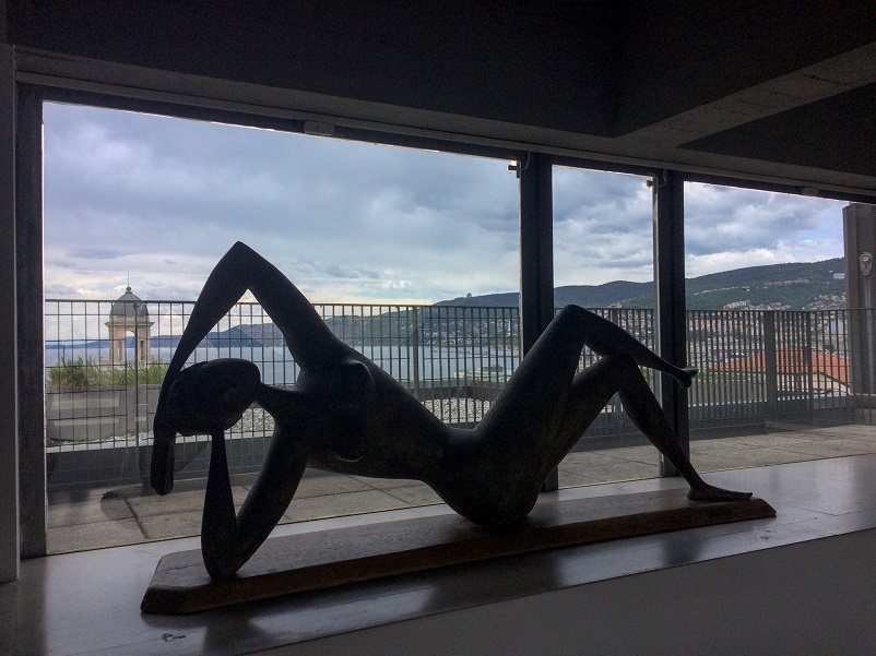 Sculpture of a reclining woman in front of a wide window onto Trieste waterfront