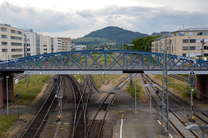 View of Wiwilibrücke - a small blue bridge over train tracks with a mountain in the back, Freiburg