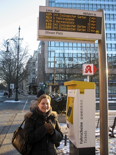 Sarah standing at a bus stop under a digital sign with arrival times in Berlin