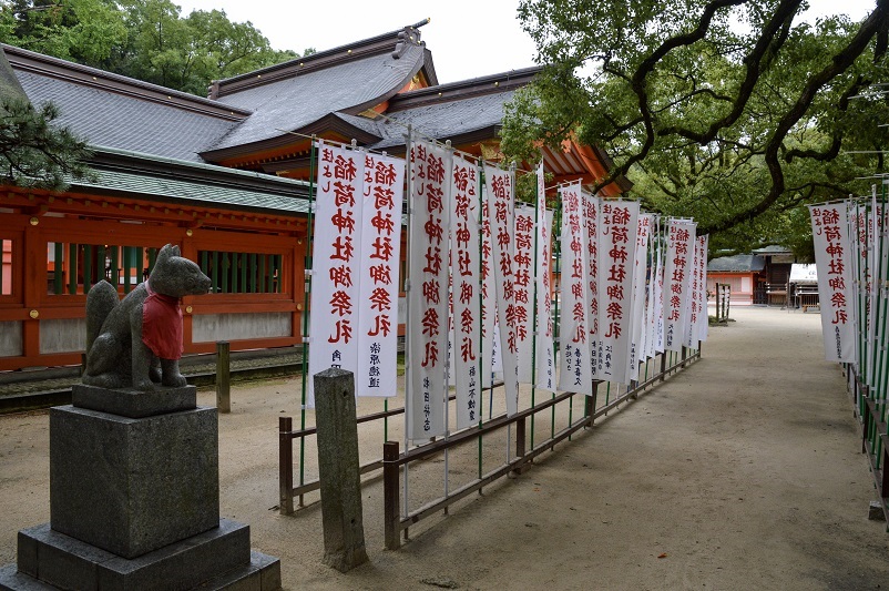 Small fox statue and flags outside an orange building at a shrine in Fukuoka