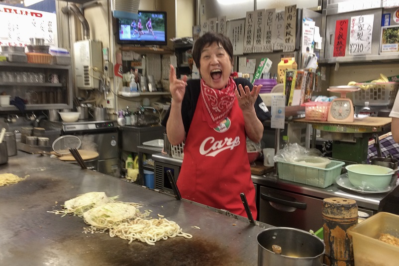 Super excited woman wearing a Hiroshima Carp apron standing in front of a flatop with okinomiyaki