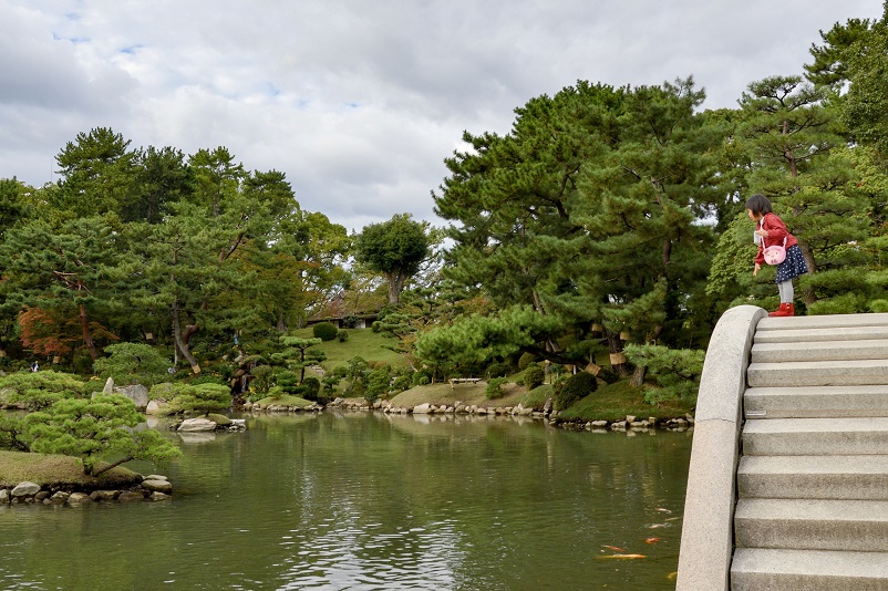 Small girl leaning off bridge to feed koi fish at Shukkei-en Garden in Hiroshima, Japan