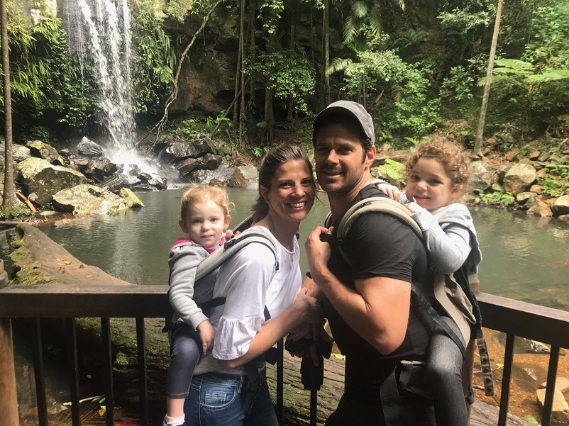 A man and a woman with two small girls on their backs standing in front of a waterfall in Australia