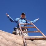 A woman standing at the top of a ladder with her arms raised against a blue sky