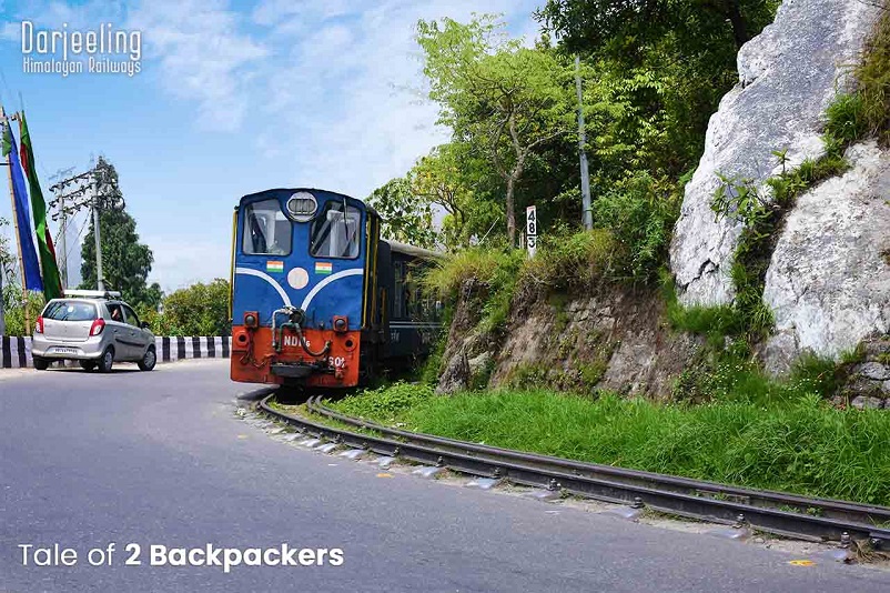 Blue train on a very small track on the side of a paved road: the Darjeeling toy train