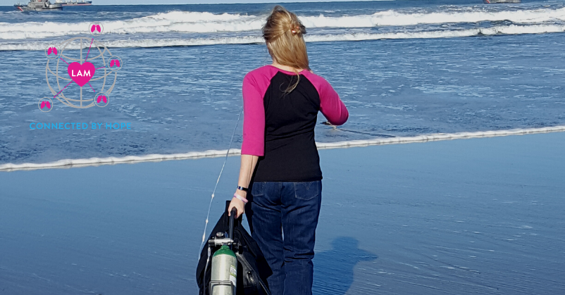 Dina Marie, a woman walking towards the ocean rolling an oxygen tank behind her; WWLAM 2020 logo in upper left corner