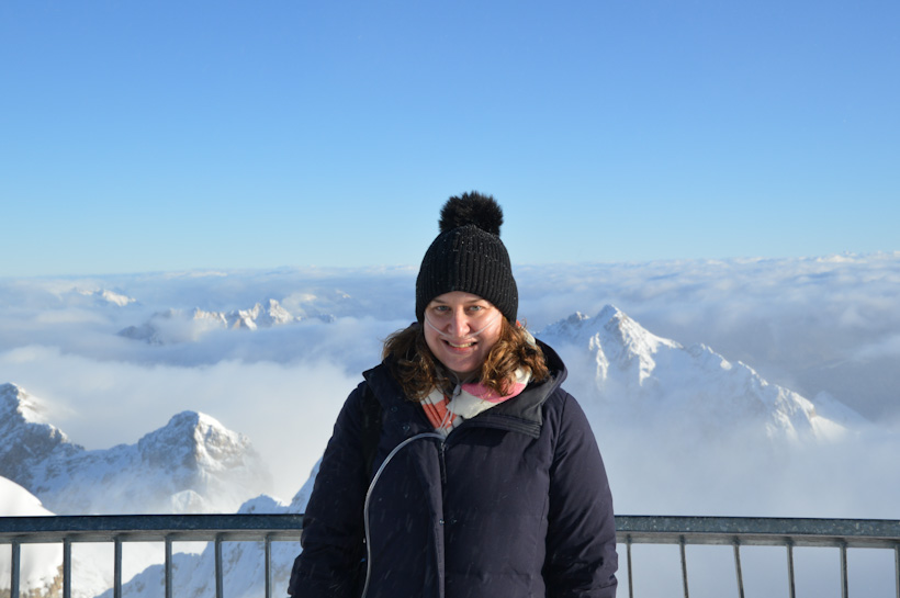 Sarah, wearing an oxygen hose, standing at the top of Germany with clouds and the tops of mountains in the background