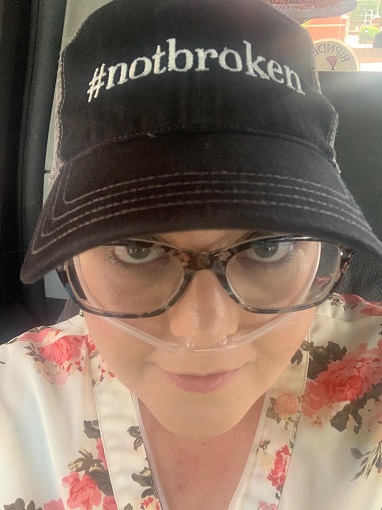 Woman wearing a black hat that says "#notbroken", also wearing glasses and an oxygen hose