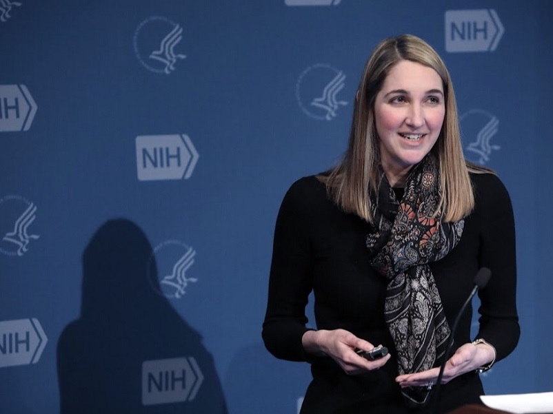 Steph speaking at the NIH, standing in front of a blue background