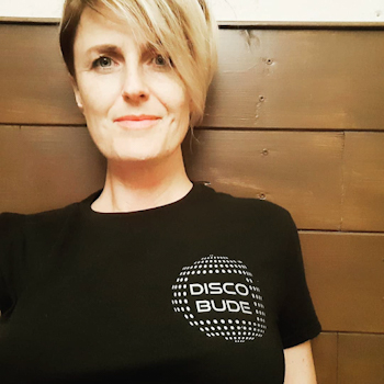 Sina: a woman with short blonde hair cut at an angle, wearing a black shirt with a small disco ball on the right side (of the photo) that reads disco bude