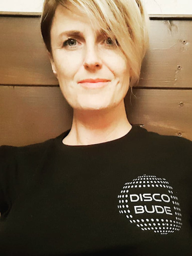 Woman with short blonde hair cut at an angle, wearing a black shirt with a small disco ball on the right side (of the photo) that reads disco bude
