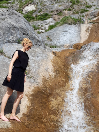 A woman, barefoot, wearing a black sleeveless dress, standing on a rocky area looking at a small waterfall