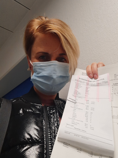 Sina (a woman with short blonde hair) wearing a mask, holding up white papers with tests results