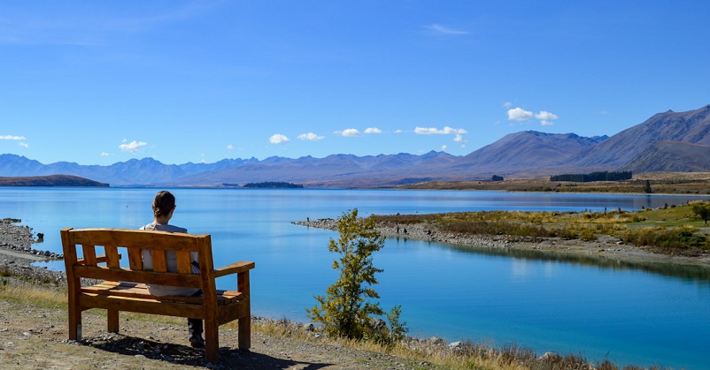 A woman sitting on a bench in front of a very blue lake and some mountains in New Zealand